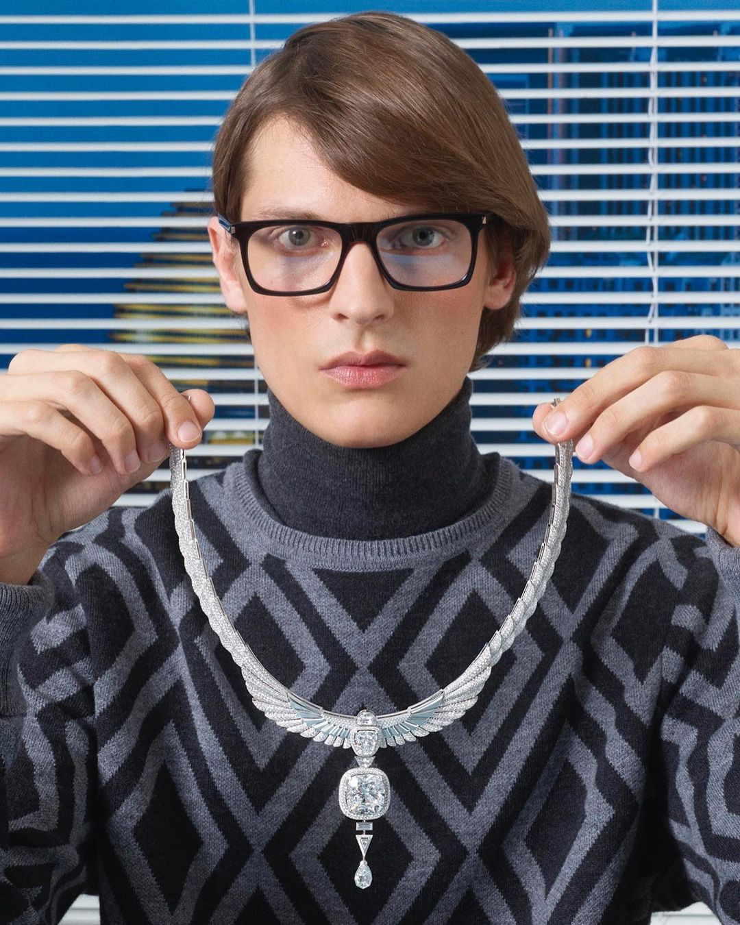 Elegant adult male showcases a silver-tone Egyptian Revival necklace indoors with natural lighting, wearing a geometric-patterned sweater and glasses.