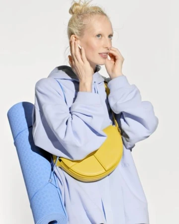 Woman in a blue hoodie holds a yellow purse and a rolled blue yoga mat against a white backdrop, wearing wireless earbuds.