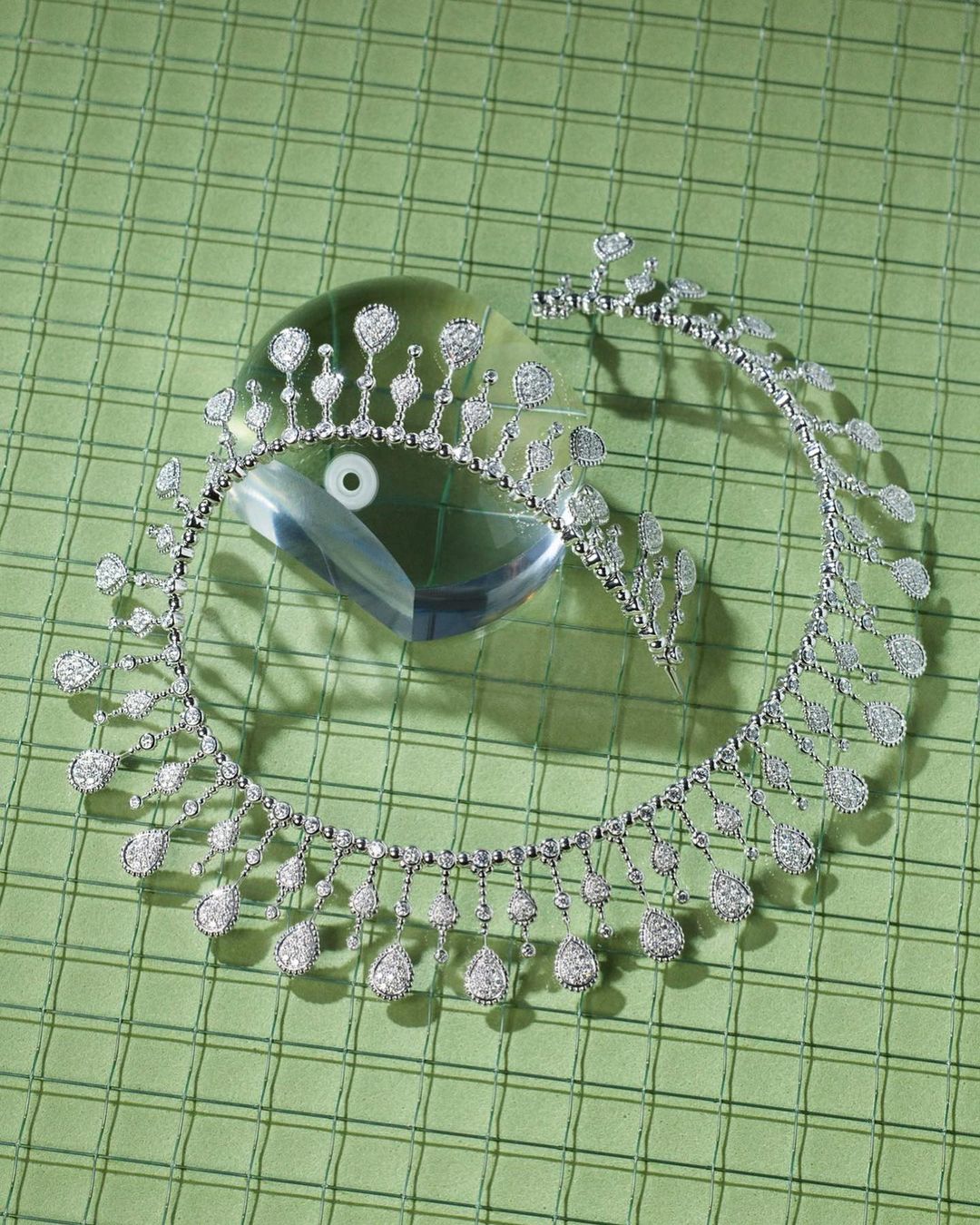 High angle studio shot of a diamond tiara and necklace displayed on a reflective crystal block atop a pale green checkered surface.