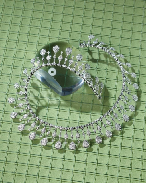High angle studio shot of a diamond tiara and necklace displayed on a reflective crystal block atop a pale green checkered surface.