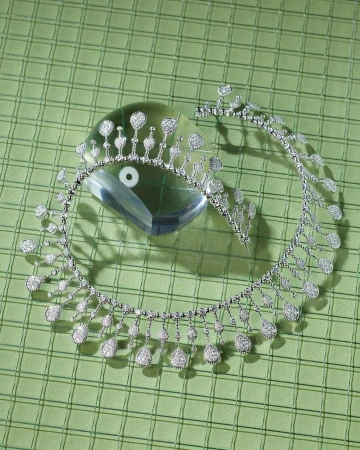 High angle studio shot of a diamond tiara and necklace displayed on a reflective crystal block atop a pale green checkered surface.