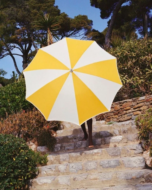 Person walks down stone steps holding yellow and white beach umbrella on a sunny day with trees and blue sky in the background.