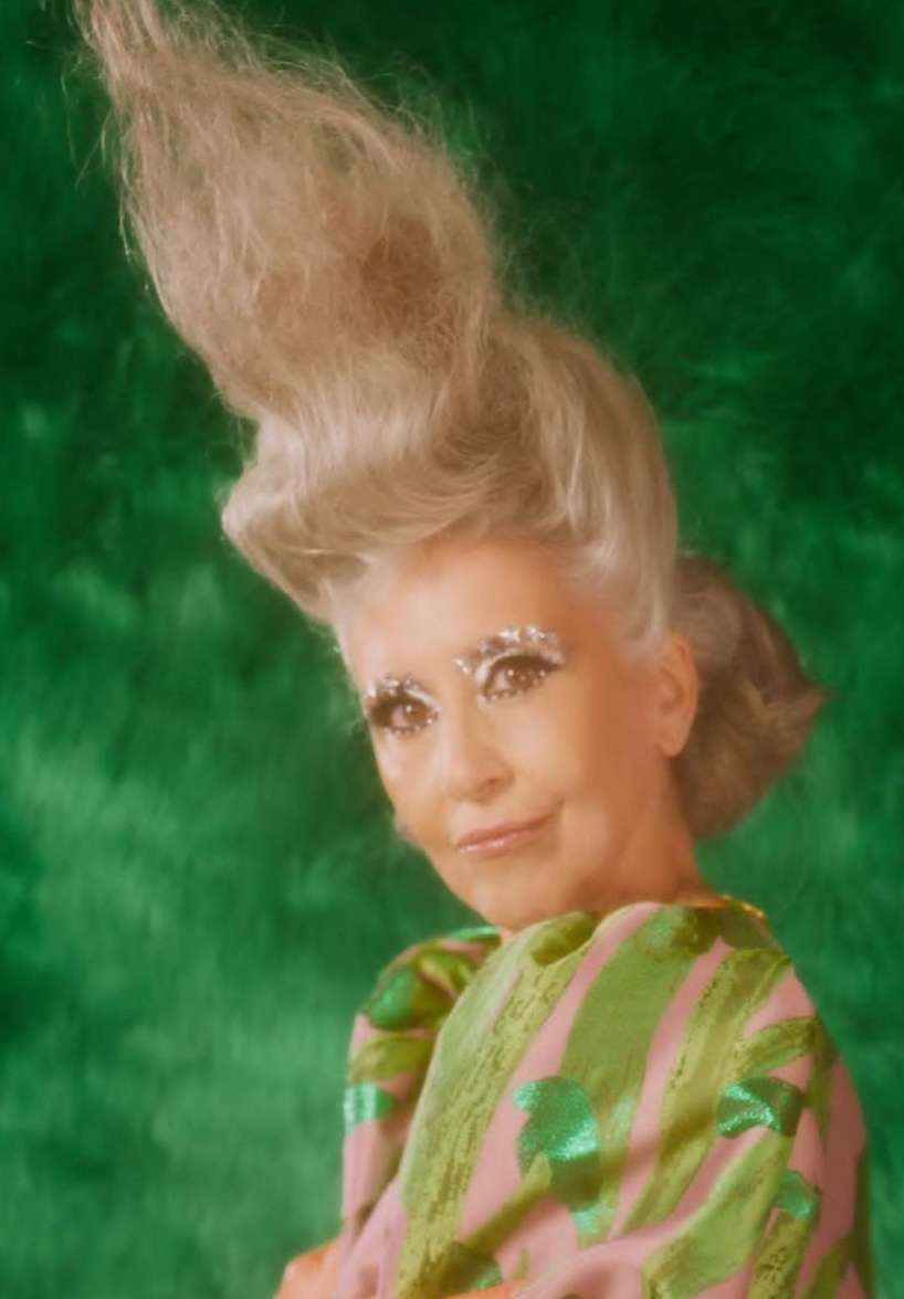 Portrait of a fair-skinned adult woman with dramatic silver makeup, light blonde updo hairstyle, and bright green backdrop and patterned top.