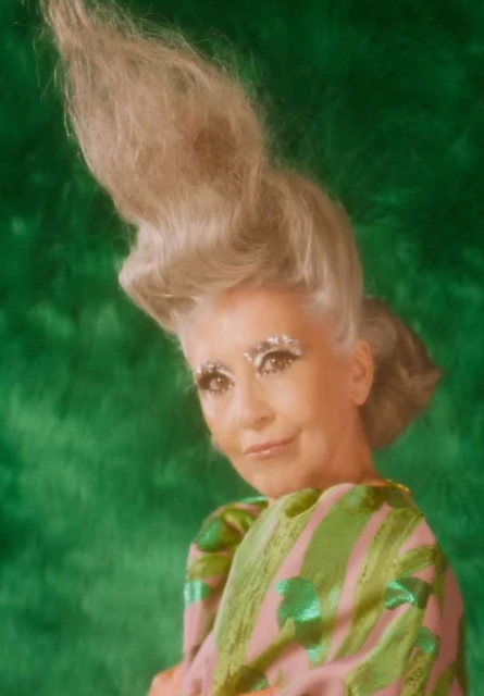 Portrait of a fair-skinned adult woman with dramatic silver makeup, light blonde updo hairstyle, and bright green backdrop and patterned top.