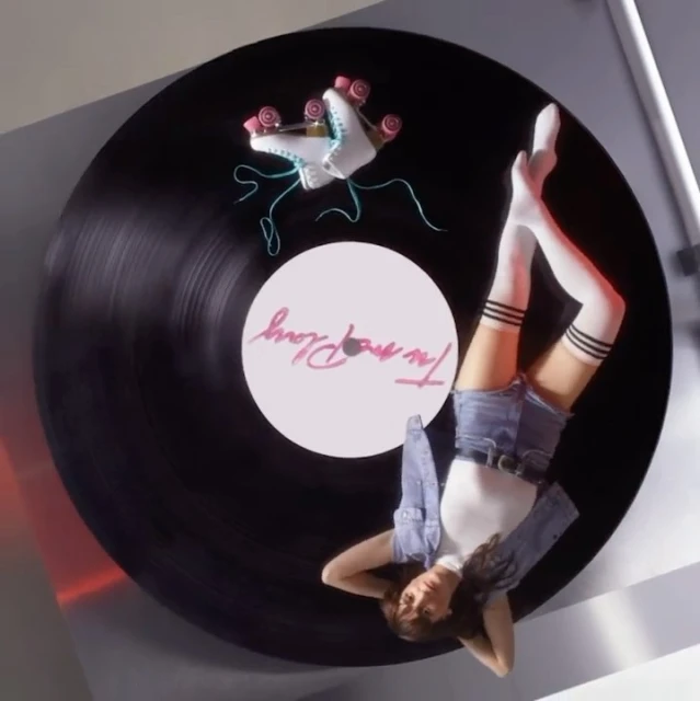 Overhead shot of a young adult woman relaxing on a black vinyl record, wearing denim shorts, white top, and roller skates nearby.