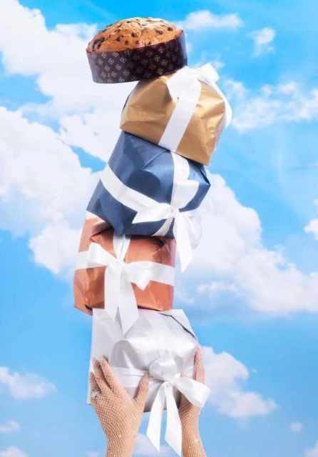 Tower of wrapped gifts and panettone is held up by a person wearing mesh gloves with a blue sky and scattered clouds background.
