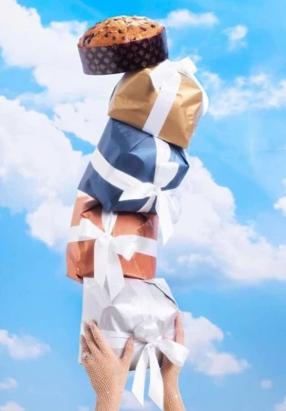 Tower of wrapped gifts and panettone is held up by a person wearing mesh gloves with a blue sky and scattered clouds background.