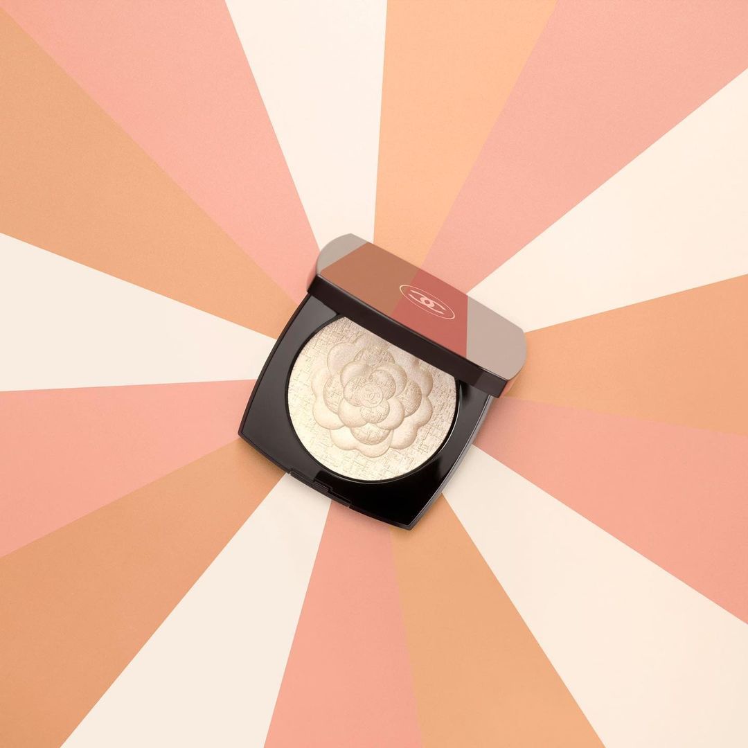 Overhead shot of Chanel illuminating powder in open black compact with flower detail, on a background of peach, orange, and cream stripes.