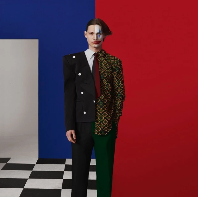 Male fashion model poses in split-design blazer with makeup against a vivid blue and red backdrop with checkered floor.