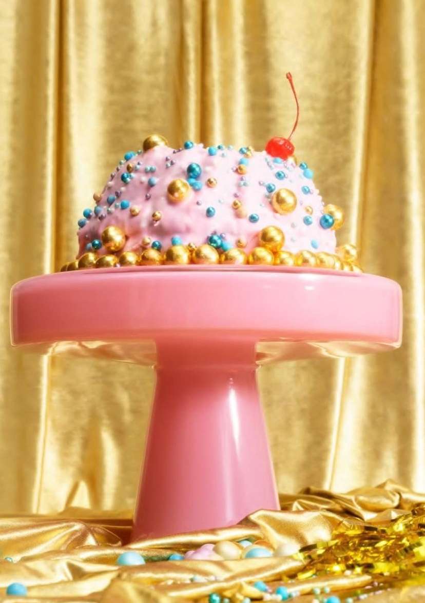 Close-up of a fanciful pink cake with gold and blue candy decorations, topped with a cherry, on a pink pedestal against a gold curtain backdrop.