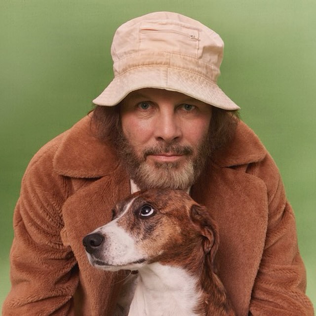 A man in a tan bucket hat and brown coat poses with a brown and white dog in front of a solid green background for a portrait.