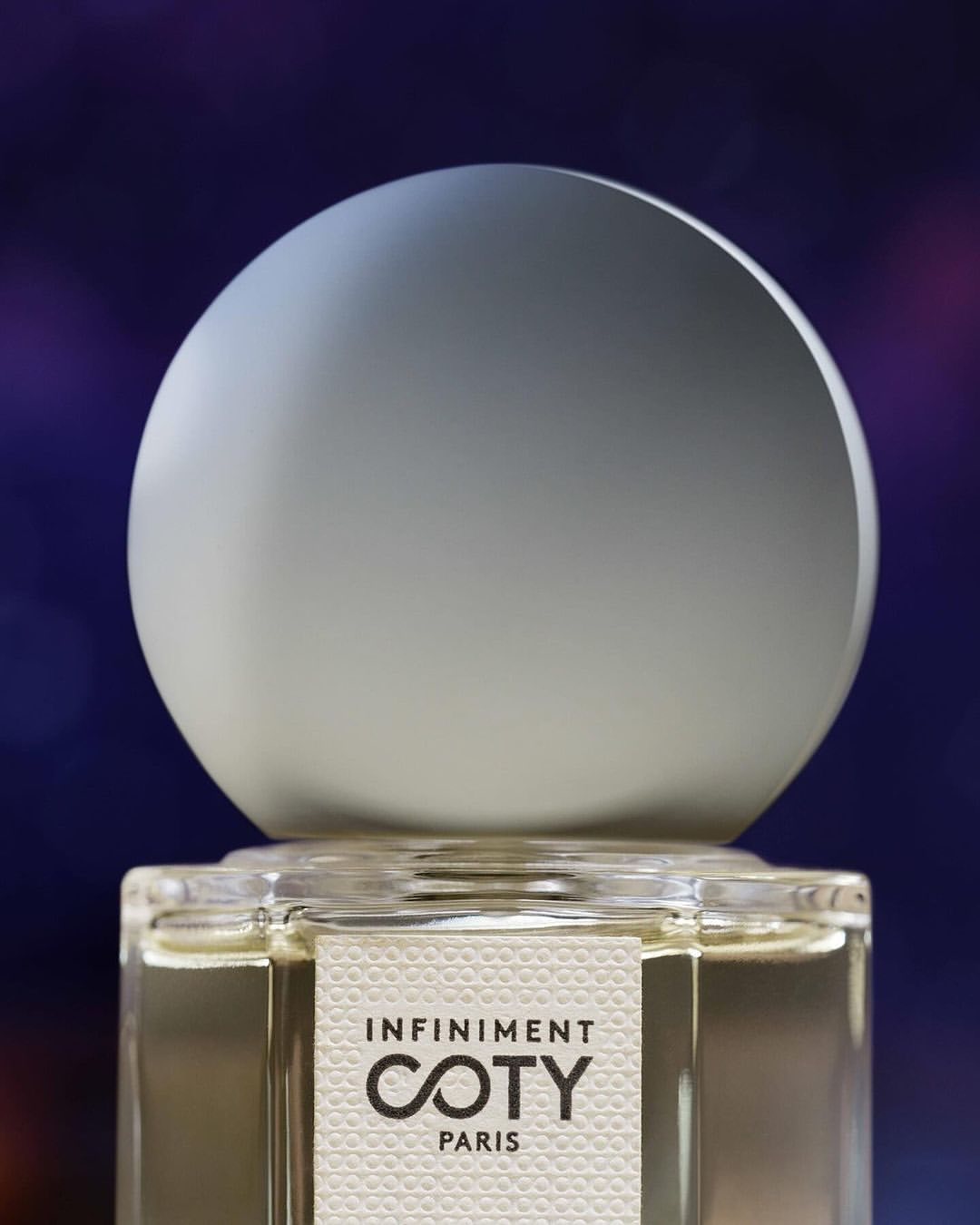 Close-up of an Infinitment Coty Paris perfume bottle with a white spherical cap against a blurred, colorful background.