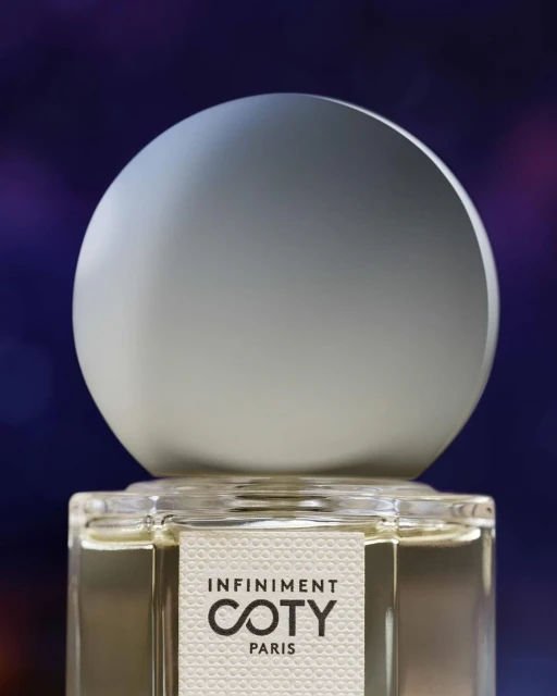 Close-up of an Infinitment Coty Paris perfume bottle with a white spherical cap against a blurred, colorful background.