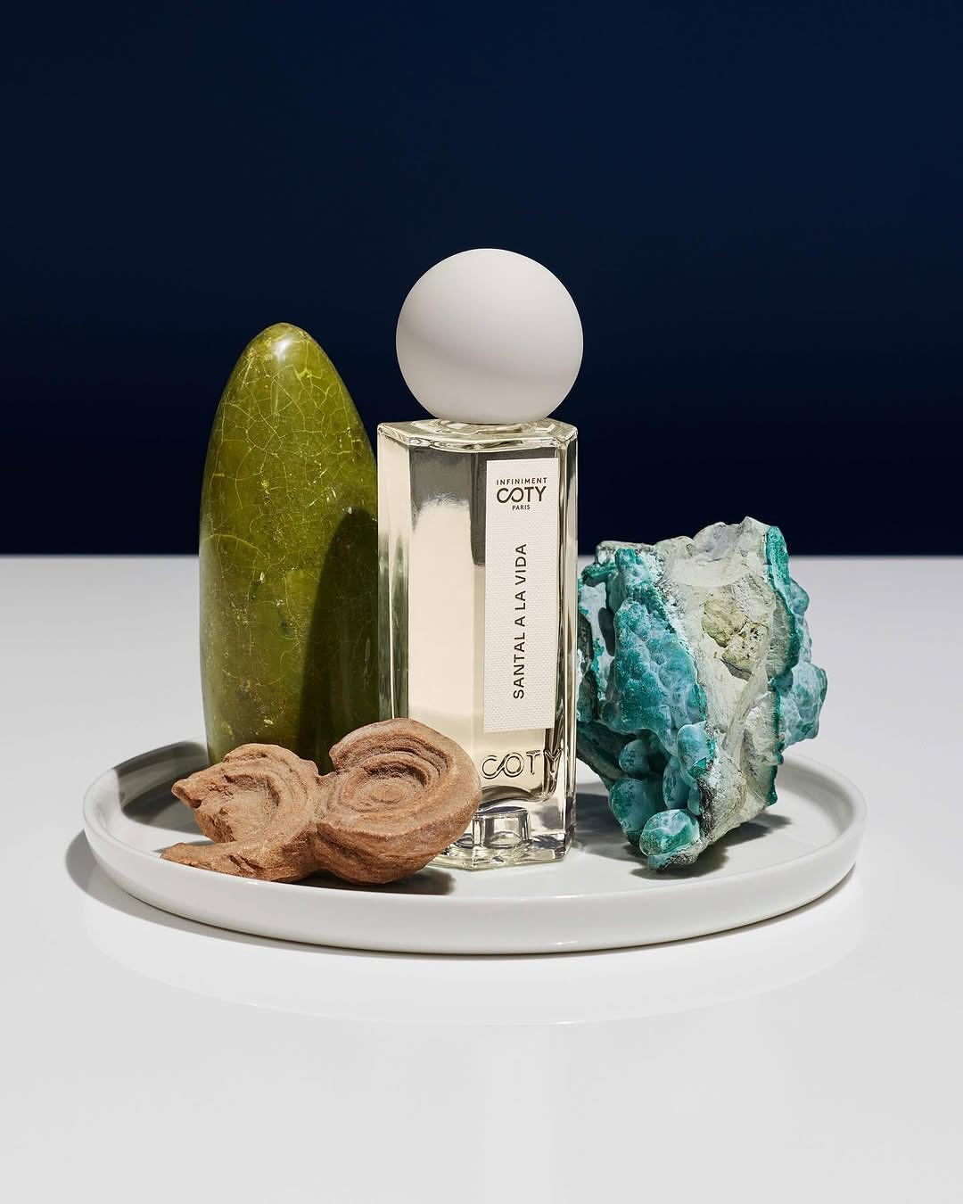 Studio shot of Coty Santal A La Vida perfume bottle on a white plate with textured stones, against a graduated navy blue background.