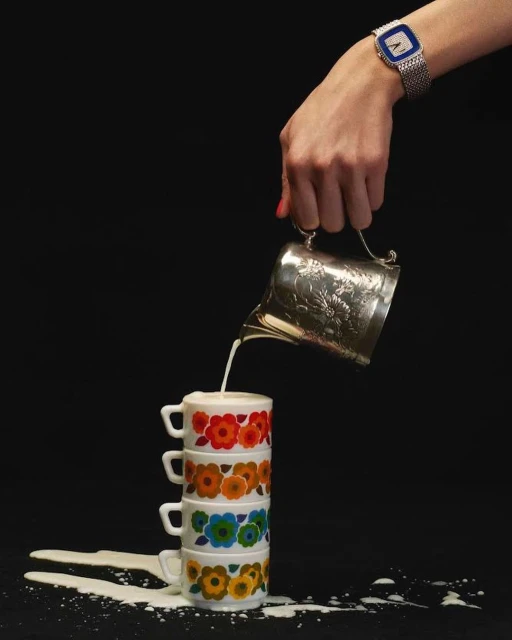 A hand pours milk from a silver creamer onto a stack of four floral patterned cups with spilled milk around the base, studio shot.