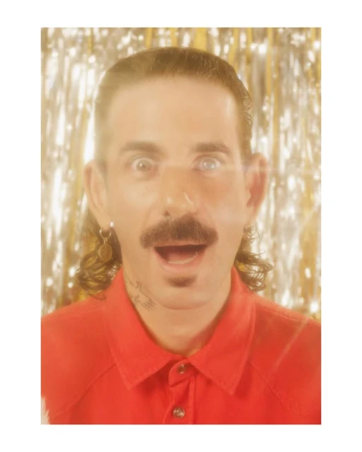 Close-up portrait of a man with a mustache and goatee, looking surprised against a gold tinsel background, wearing a red button-up shirt.