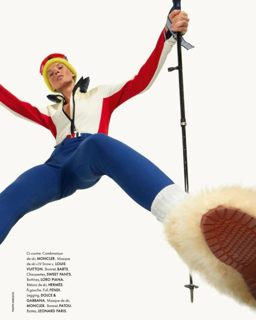 Fashion editorial shot of a model in a ski suit and hat, holding ski poles against a white backdrop, emphasizing clothing and pose.