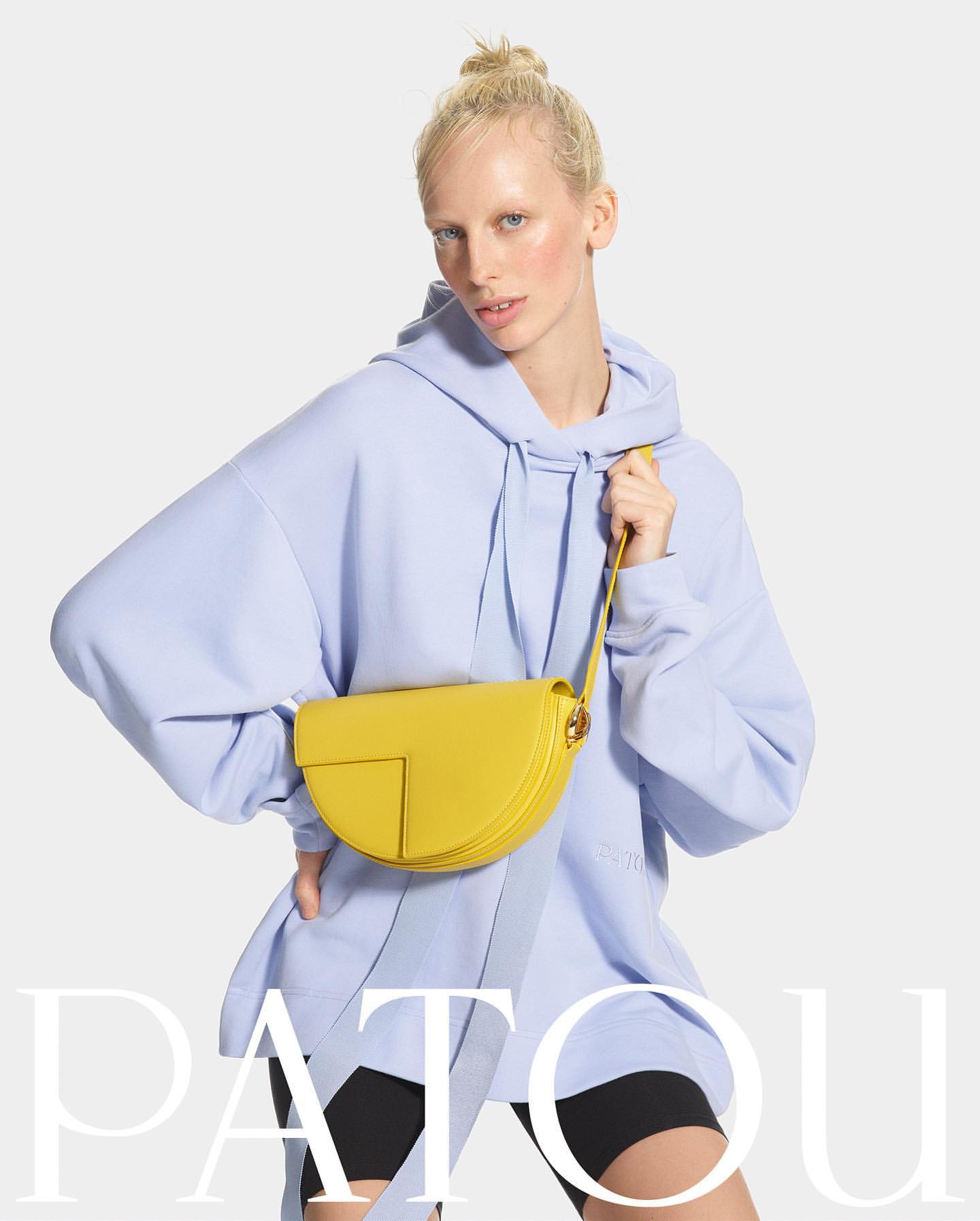 Blonde model in blue hoodie holding yellow Patou handbag, posing in a studio setting with a neutral background and visible brand name.