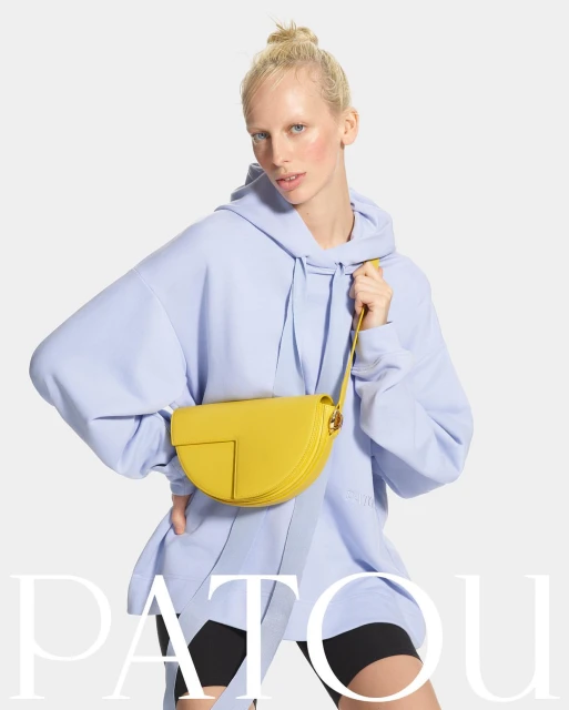 Blonde model in blue hoodie holding yellow Patou handbag, posing in a studio setting with a neutral background and visible brand name.
