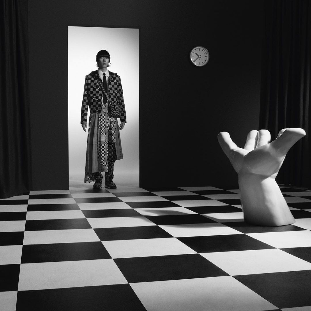 Monochrome fashion shot: An adult male poses in a checkered suit and long skirt, standing in a doorway in a surreal room with a checkered floor.