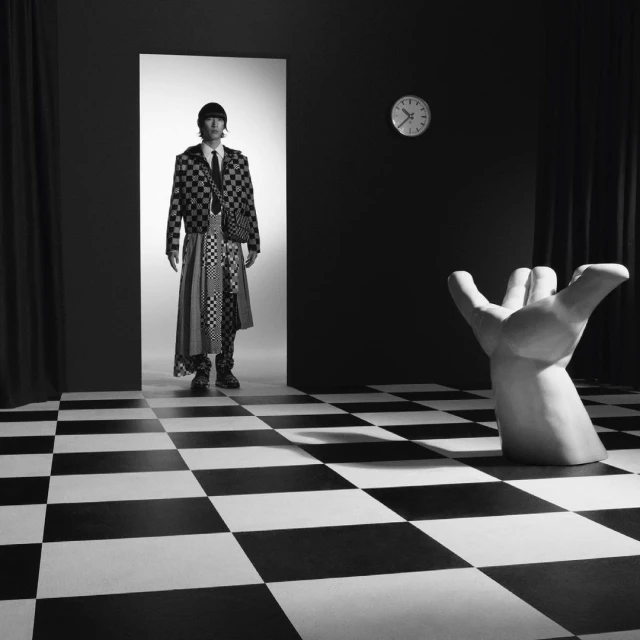 Monochrome fashion shot: An adult male poses in a checkered suit and long skirt, standing in a doorway in a surreal room with a checkered floor.