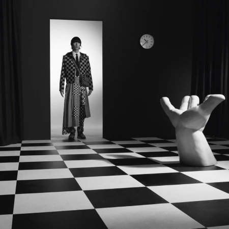 Monochrome fashion shot: An adult male poses in a checkered suit and long skirt, standing in a doorway in a surreal room with a checkered floor.