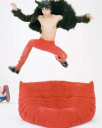 Shirtless man mid-air leaping over a red tufted sofa wearing a dark fur jacket and bright red trousers