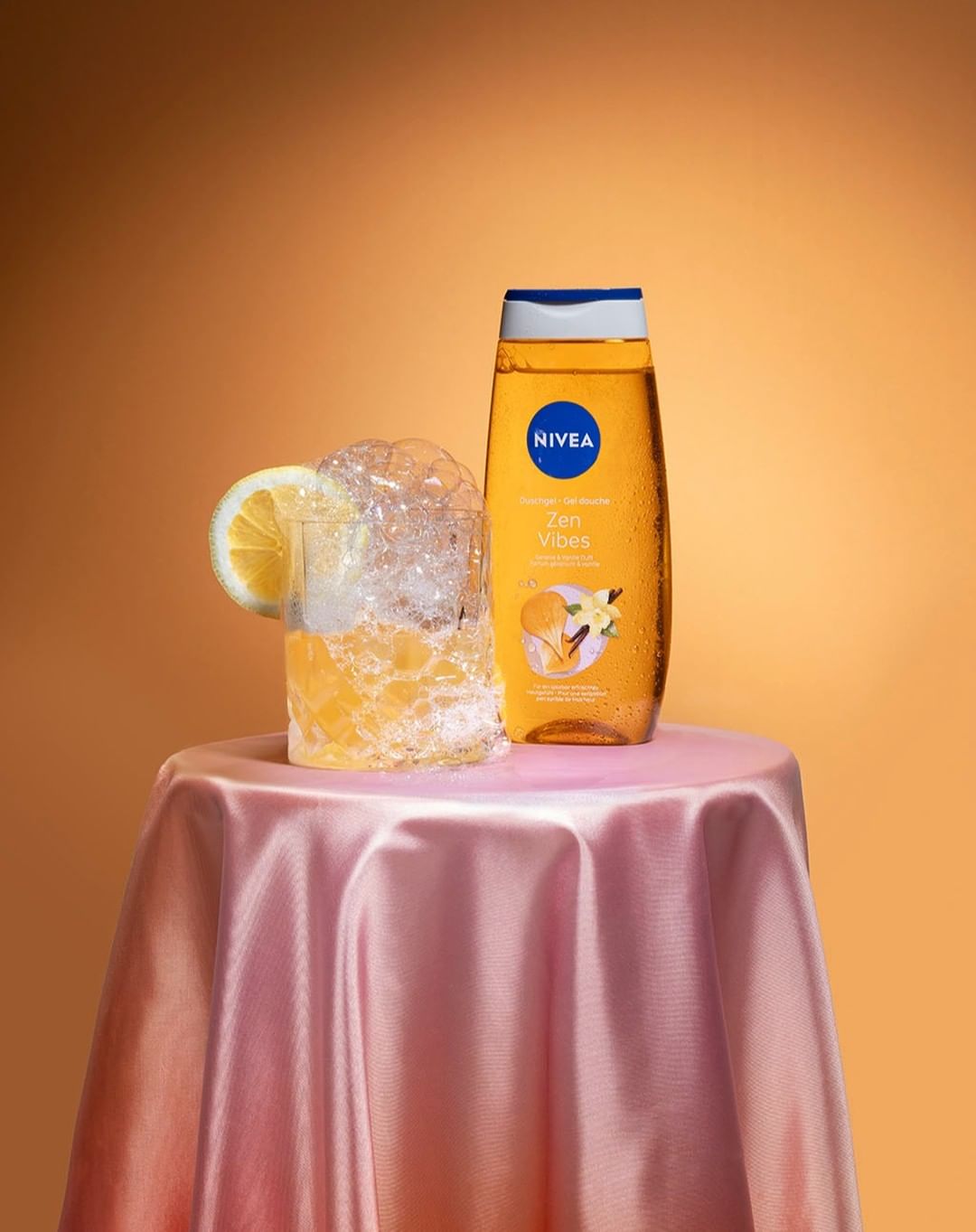 Still life shot of Nivea shower gel bottle next to glass with lemon and bubbles, on a pink silk draped table against an orange background.