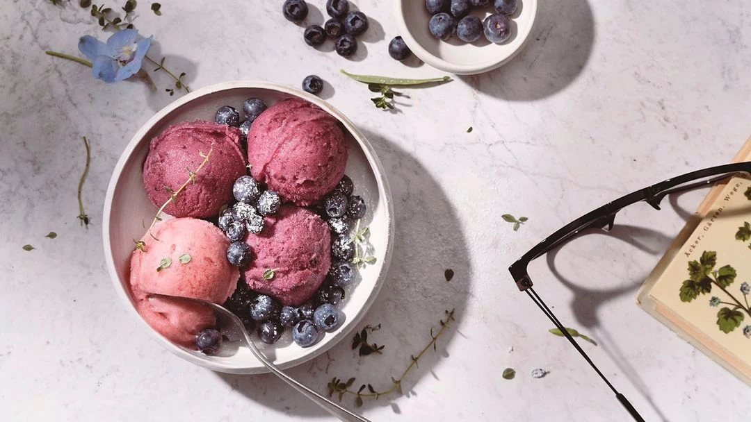 Still life featuring scoops of fruit sorbet in a bowl, garnished with blueberries and herbs, next to a book and eyeglasses on marble.