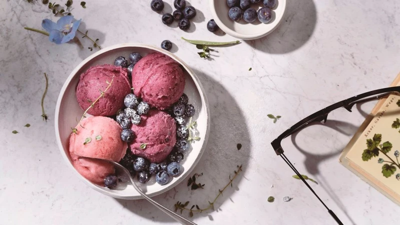 Still life featuring scoops of fruit sorbet in a bowl, garnished with blueberries and herbs, next to a book and eyeglasses on marble.