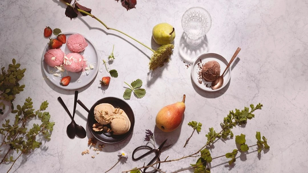 Overhead shot of plates of ice cream, pears, strawberries, flowers, and greenery on a marble surface with copper and black spoons.