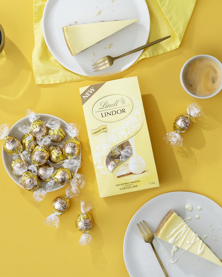 Overhead shot of cheesecake slices, Lindt Lindor truffles, coffee cup, and gold forks on a vibrant yellow surface.