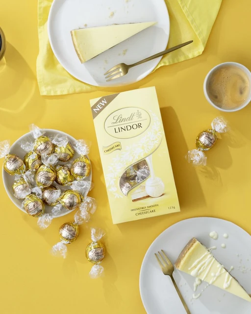 Overhead shot of cheesecake slices, Lindt Lindor truffles, coffee cup, and gold forks on a vibrant yellow surface.
