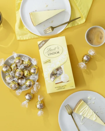 Overhead shot of cheesecake slices, Lindt Lindor truffles, coffee cup, and gold forks on a vibrant yellow surface.