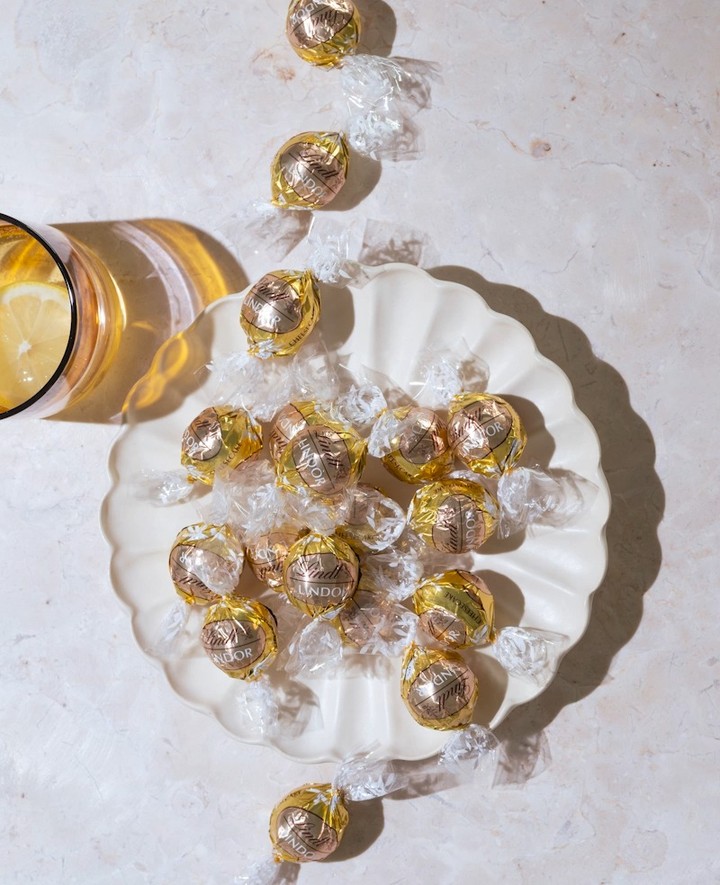 Overhead shot of gold-foil-wrapped Lindor chocolates arranged on a decorative white plate, next to a golden-tinted glass of lemonade.