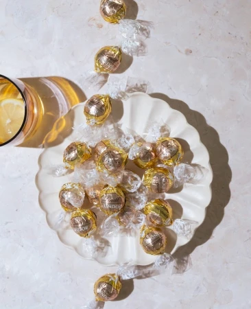 Overhead shot of gold-foil-wrapped Lindor chocolates arranged on a decorative white plate, next to a golden-tinted glass of lemonade.