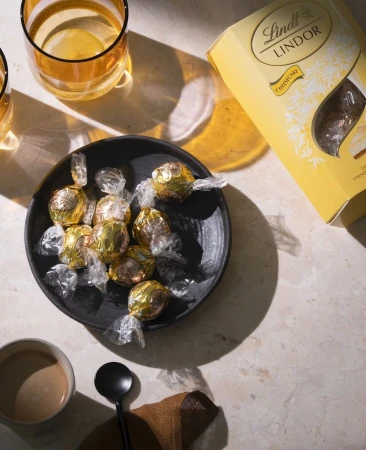 Overhead shot of Lindt Lindor chocolates in a black bowl with glasses and a cup of coffee on a marble surface.