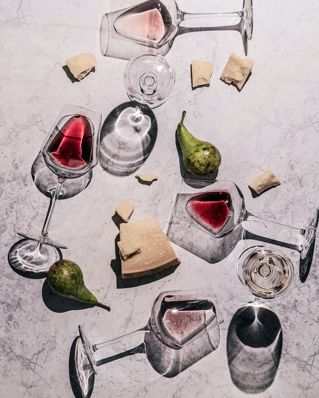 Overhead shot of red and white wine in glasses with pears and parmesan cheese on a textured light gray surface.