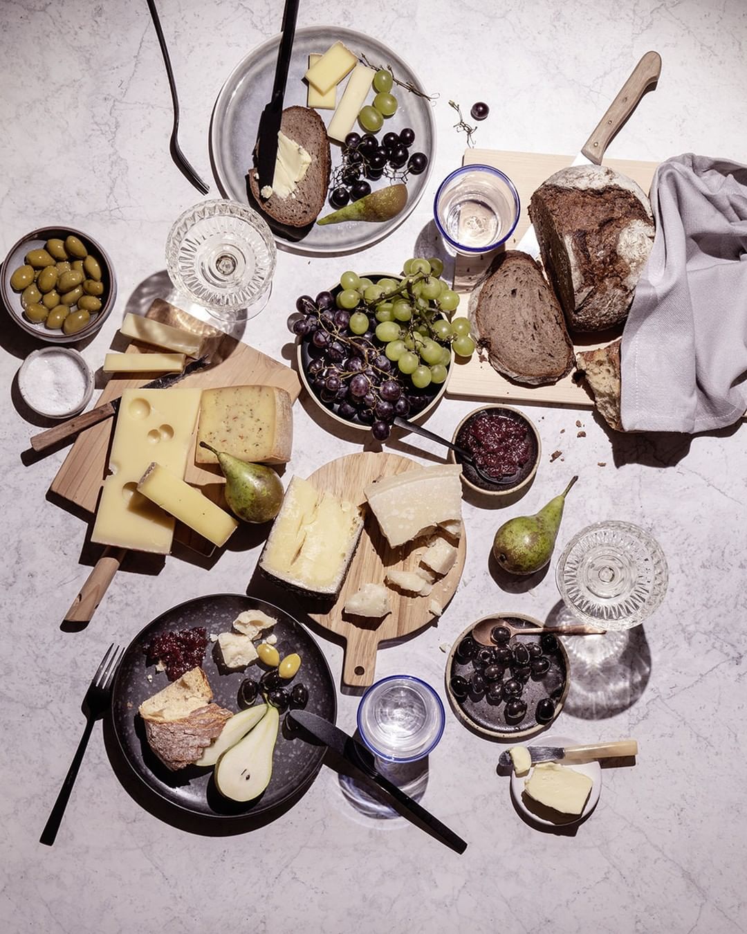 Overhead shot of cheese, bread, grapes, olives, jam, water glasses, and cutlery on a marble surface, creating a delicious food still life.