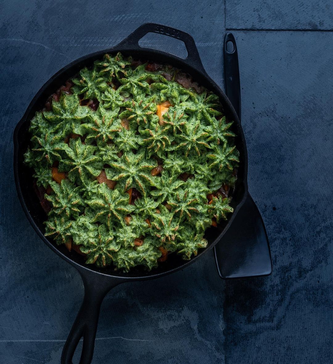 Overhead shot of shepherd's pie in cast iron skillet with green piped topping, next to spatula on dark blue textured surface.