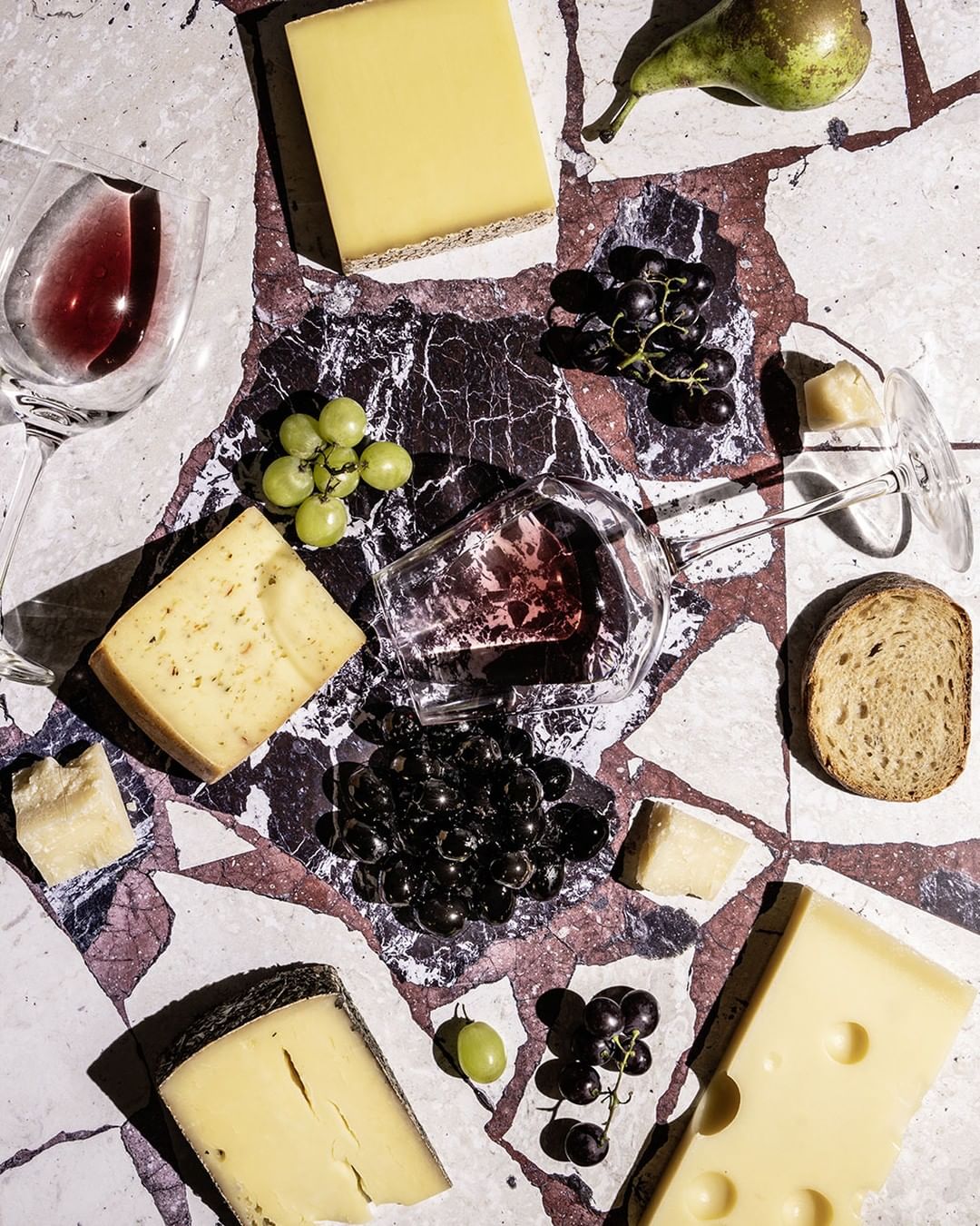 Overhead shot of cheese slices, grapes, bread, pear, and glasses of red wine on a marble surface creating a rustic food still life.