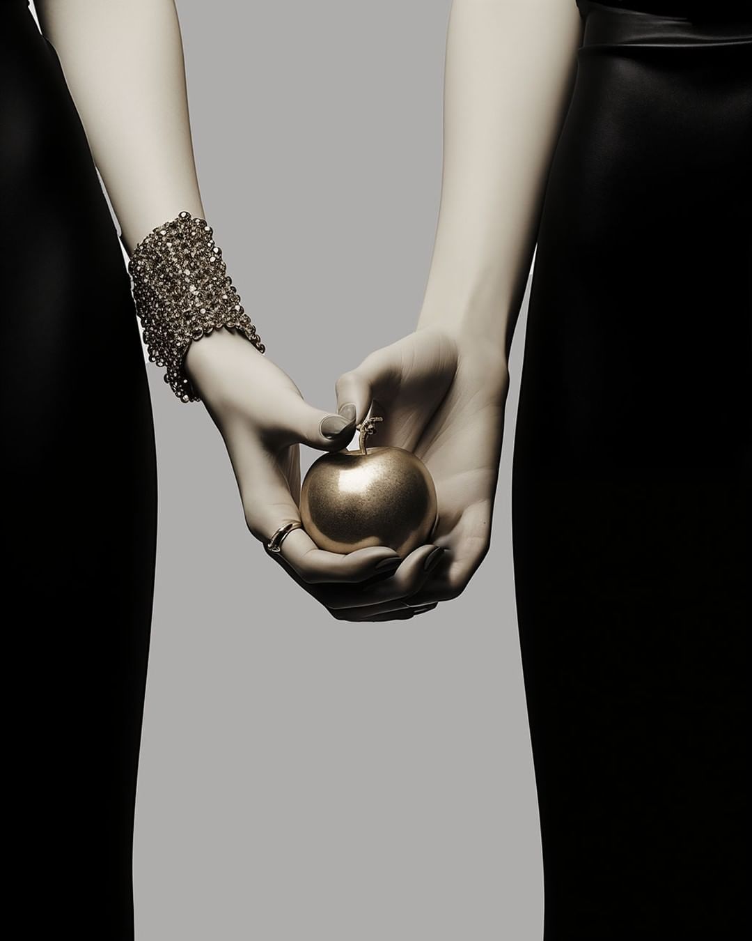 Two figures hold a gold apple against a gray backdrop, one arm adorned with a mesh bracelet, bodies only partially visible in black attire.