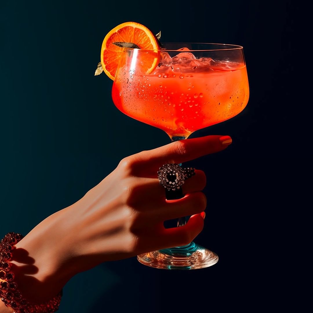 Woman's hand holds a cocktail glass filled with a red drink, garnished with an orange slice against a dark teal background.