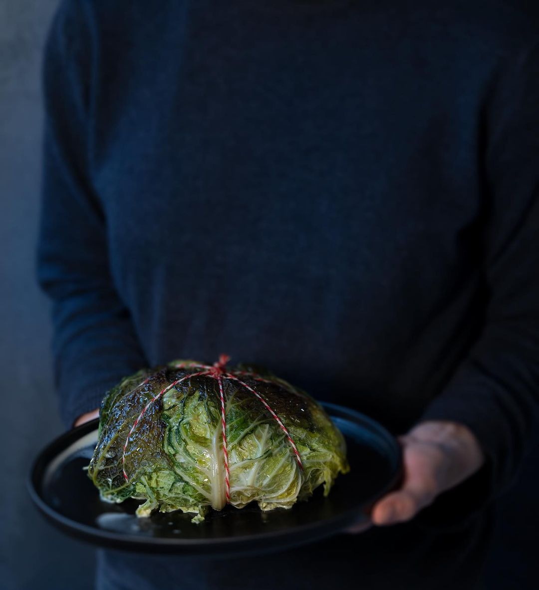 A person holds a black plate with a cabbage wrapped in string, highlighting culinary presentation and food photography against a gray backdrop.