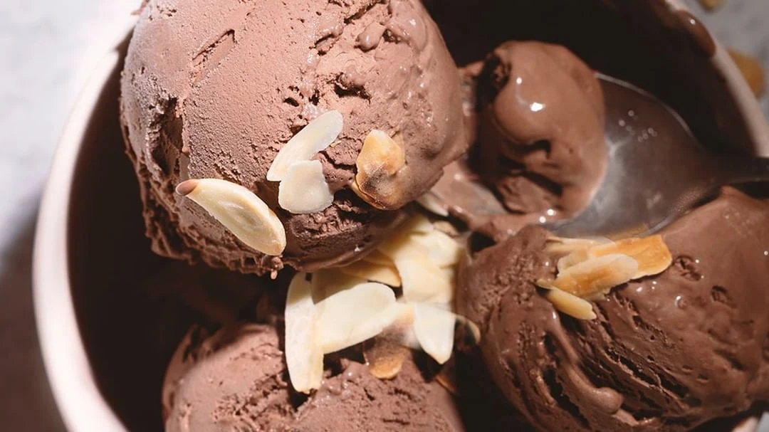Bowl filled with scoops of rich chocolate ice cream, generously topped with sliced almonds, under a soft, diffused light.