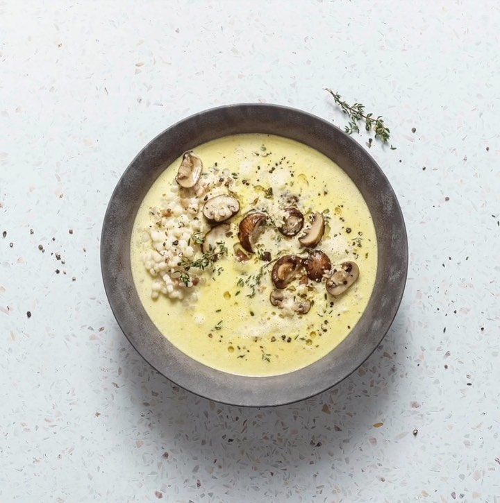 Overhead shot of a bowl of creamy soup with mushrooms, herbs, and pearl couscous on a textured white surface, food photography.