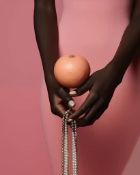 Close-up of a person with dark skin holding a round grapefruit and pearl strands against a pink background.