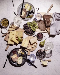 Overhead flat lay of an abundant charcuterie and cheese board with grapes, pears, bread, and wine glasses on a marble surface.