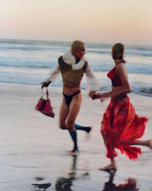Two people running on a beach with the ocean and sky in the background, one person holding a purse.