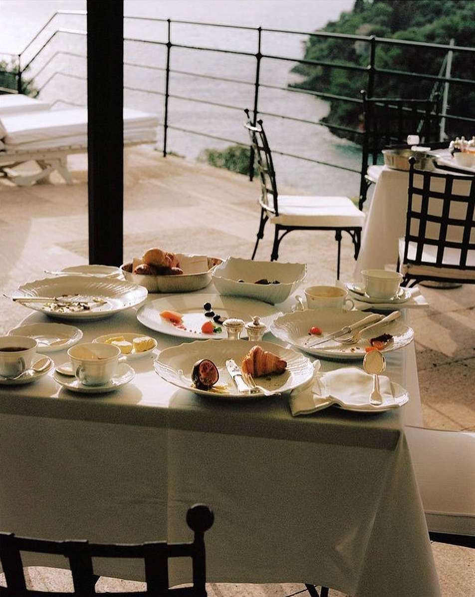 Outdoor breakfast table setting with croissants, fruit, coffee, and ornate white porcelain on a veranda overlooking a body of water.